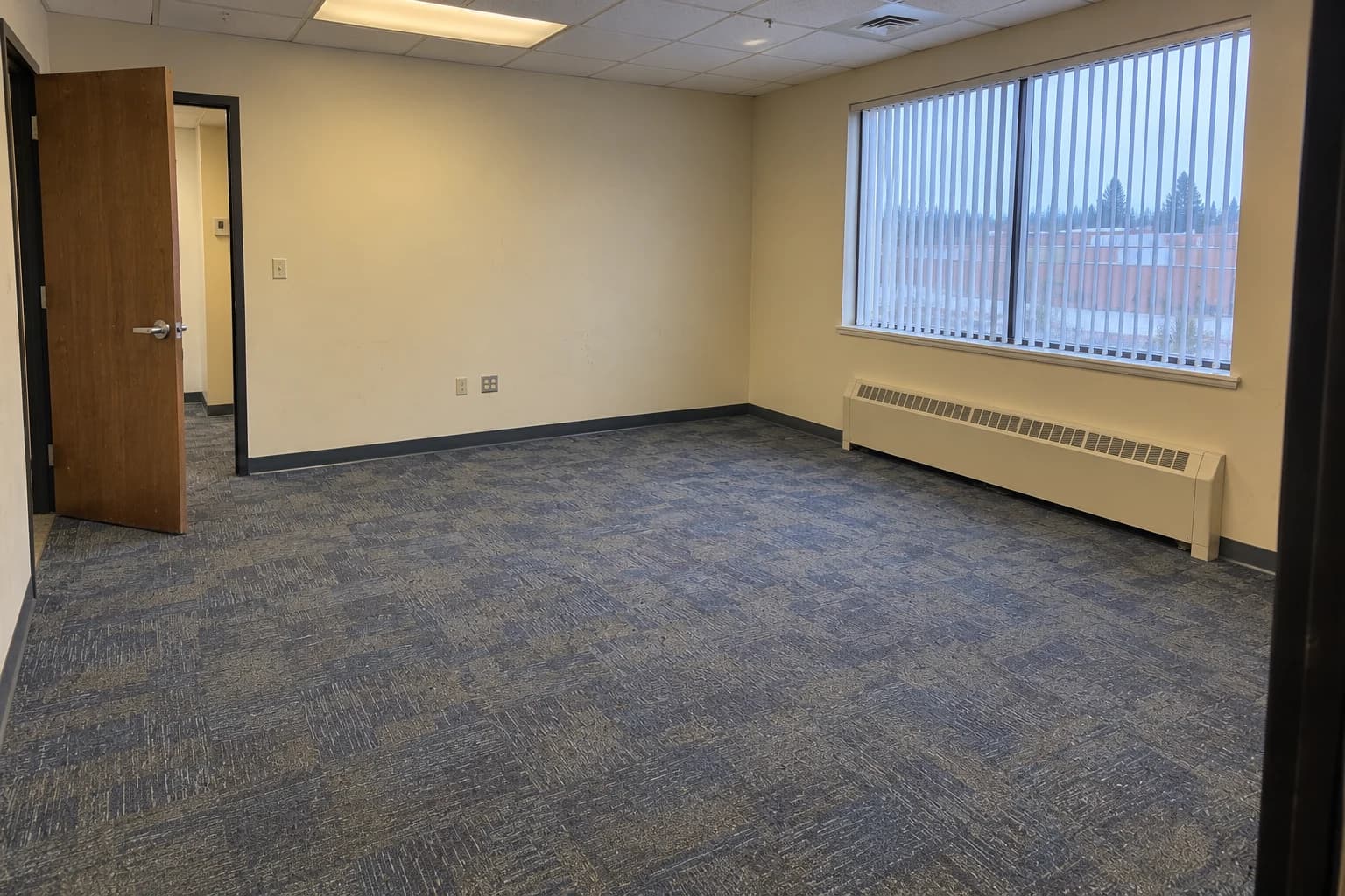 Generated after example: the same office photographed from the opposite doorway, looking back toward the entrance with the window on the right and the worn carpet unchanged